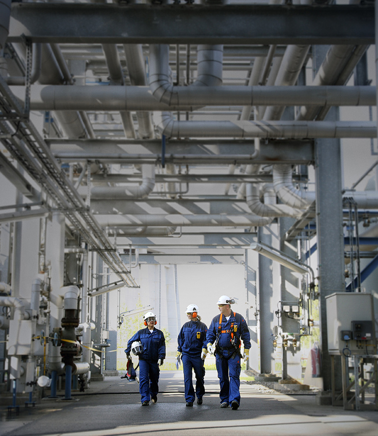 Three workers in protective gear walking through an industrial plant with overhead piping